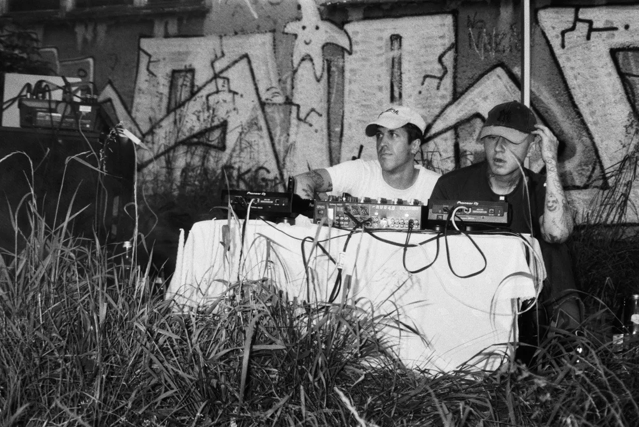 Two DJs performing behind a low booth on a grass floor, captured in black and white.