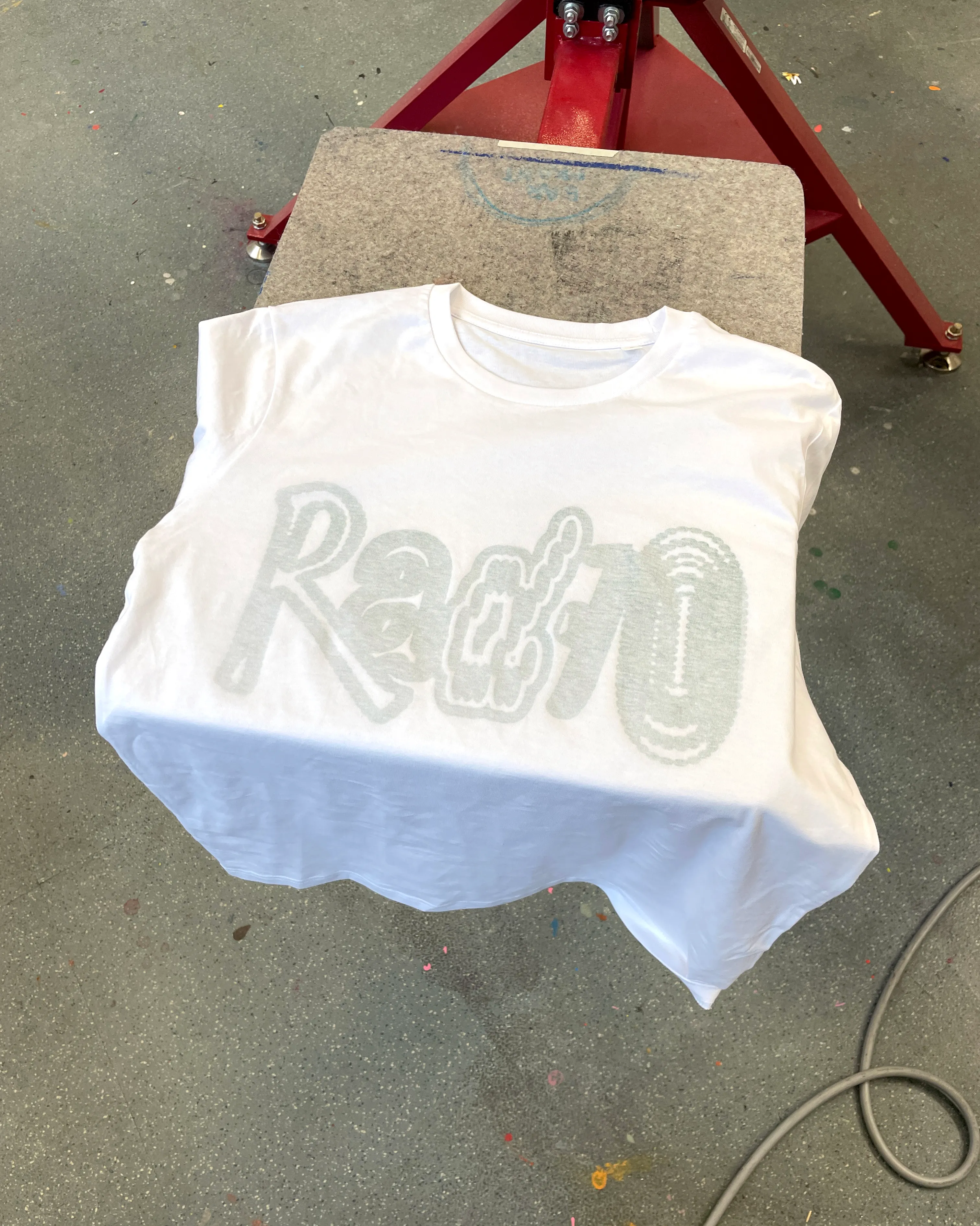 White t-shirt positioned on a screen printing press, displaying the subtle grey 'Radio' lettering printed on the inside of the fabric.